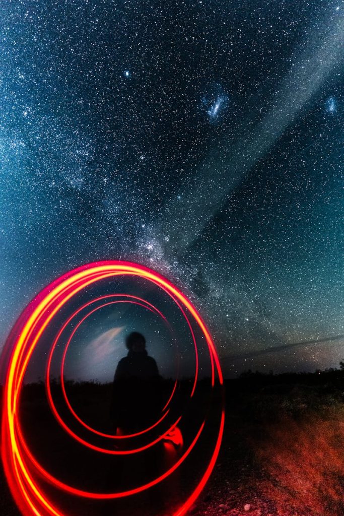 A silhouette under a starry night sky with vibrant red light trails and distant galaxies.